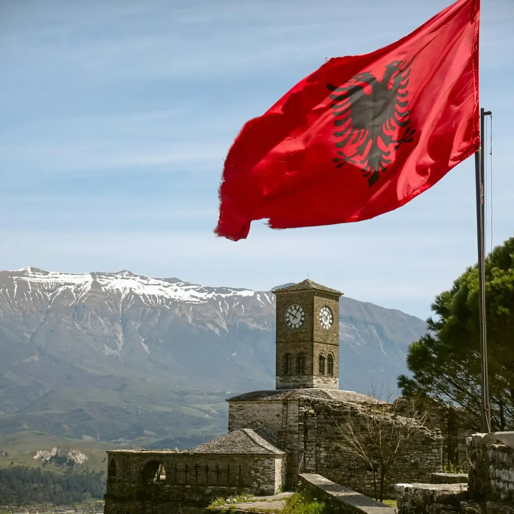 Iconic view of Gjirokaster Fortress featuring the Albanian flag and a clock tower under a blue sky.
